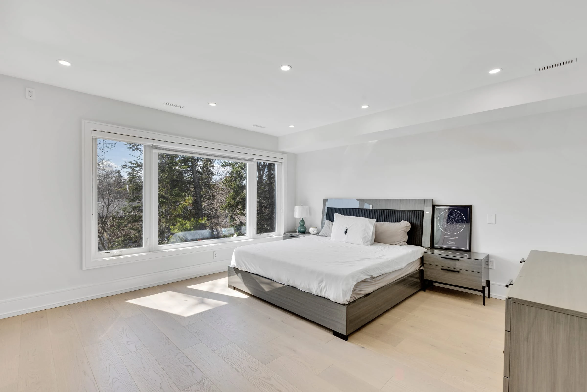 Bright primary bedroom with a tufted headboard and an oversized window over mature trees - RCG Canada